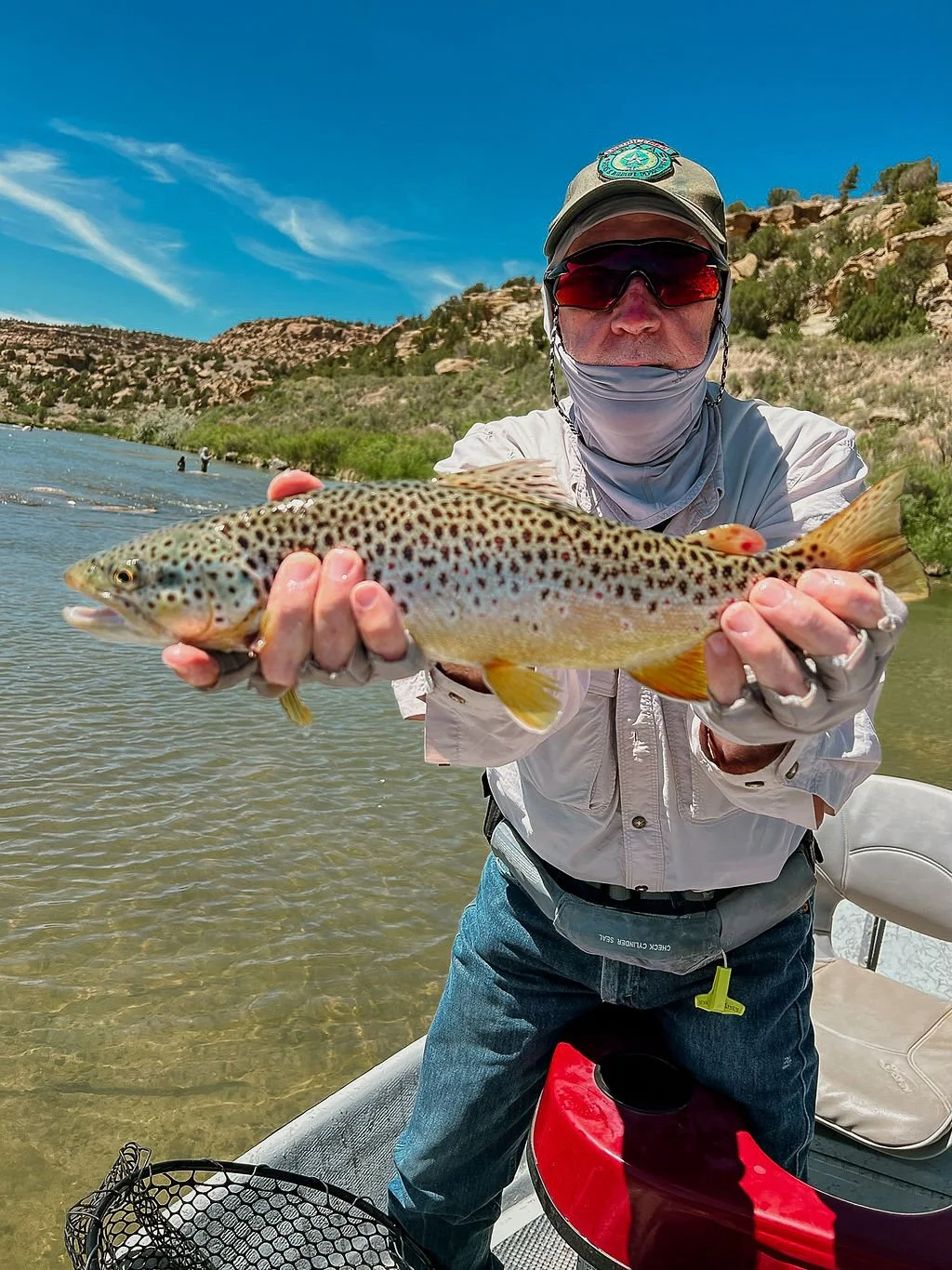 A man wearing sunglasses and outdoor clothing holding a large rainbow trout over a river with rocky hills in the background.