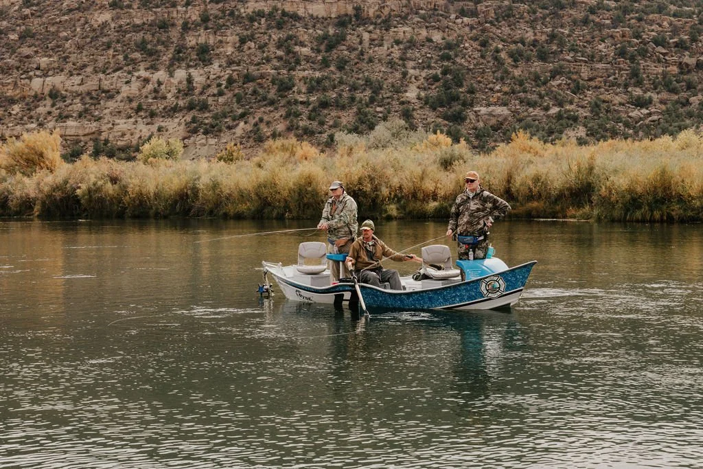 Three men fishing from a small boat on a calm river with autumn-colored bushes and a rocky hillside in the background.