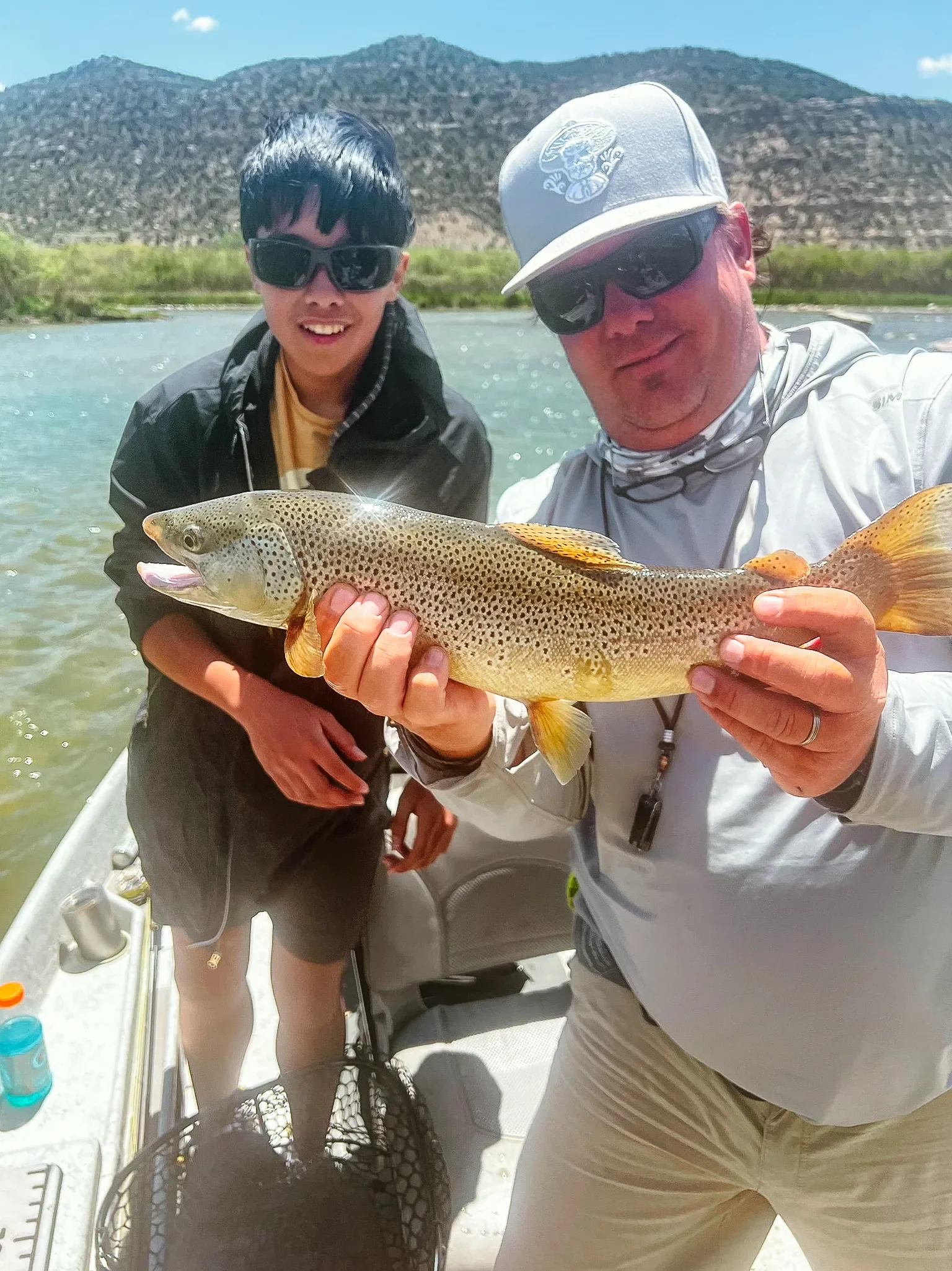 Two men holding a large fish on a boat, with mountains and water in the background on a sunny day.
