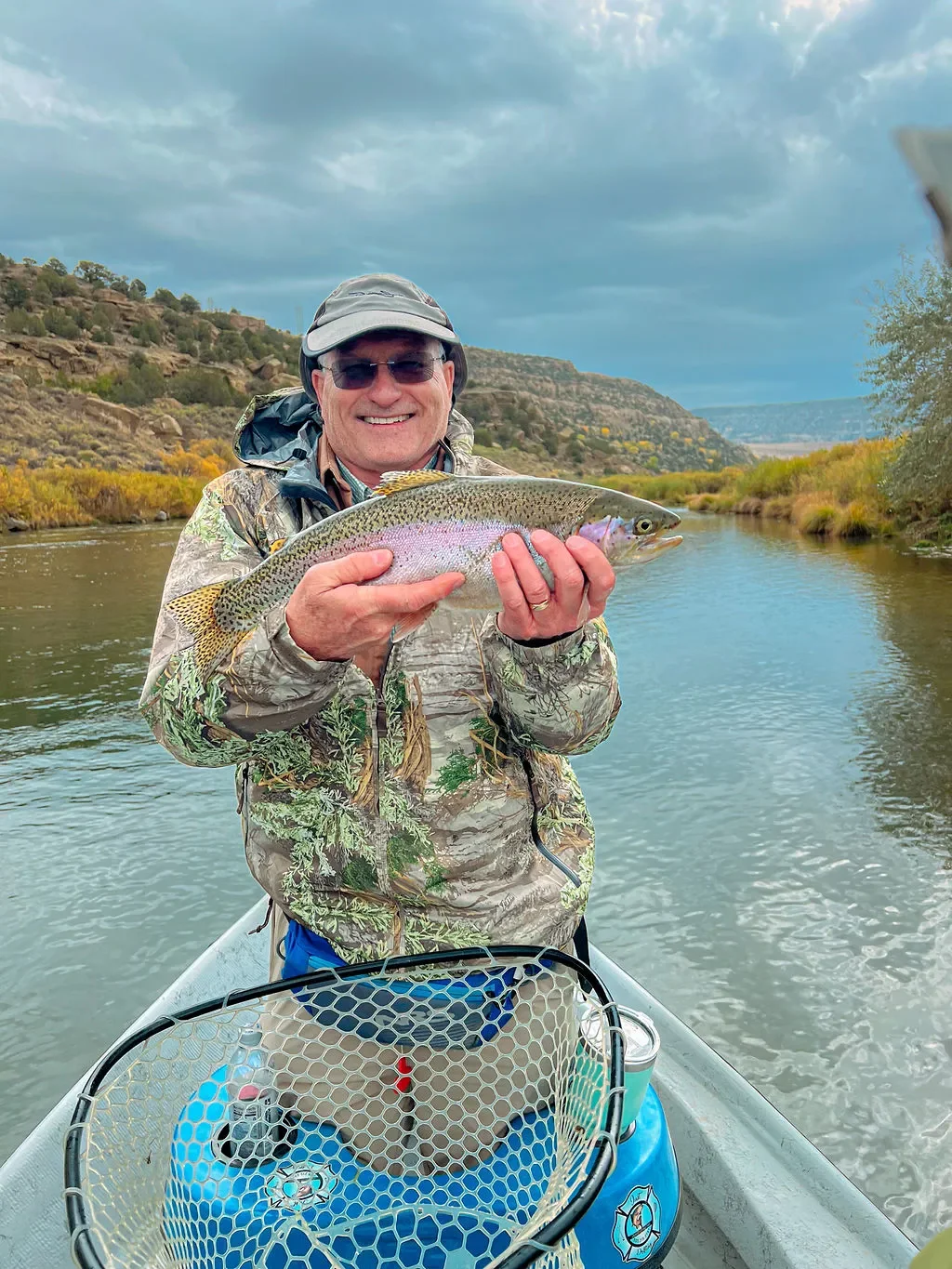 Man in camouflage jacket and baseball cap holding a rainbow trout on a boat on a river with a rocky hillside and cloudy sky in the background.