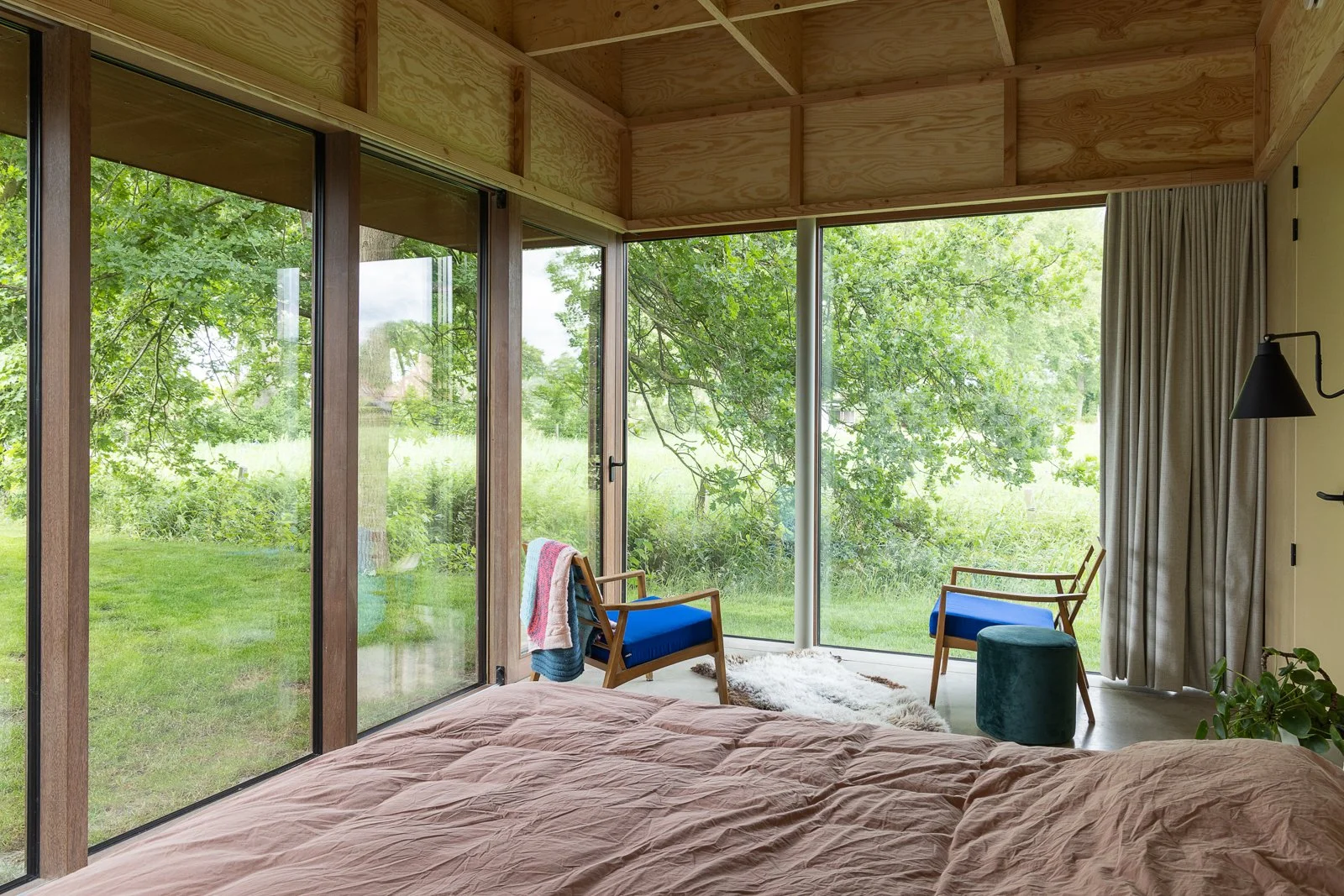 Bedroom with large glass windows overlooking trees and greenery outside. The room features a bed with pink bedding, two wooden chairs with blue cushions, a white fluffy rug, a green pouf, and cream-colored curtains.