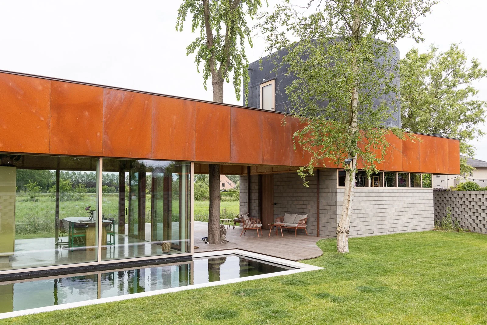 Modern house with large glass windows and an orange upper exterior, set in a grassy yard with trees and outdoor seating.
