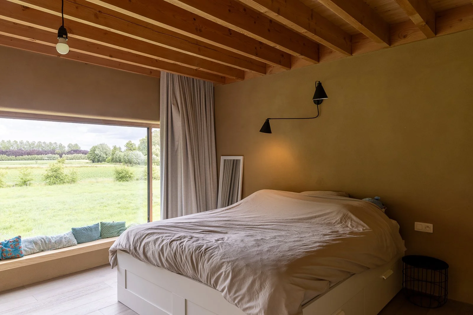 A bedroom with a large window overlooking a green field and trees. The bed has beige sheets and is positioned near the window. There are pillows on the window seat and a mirror leaning against the wall. The ceiling is wooden with exposed beams, and t