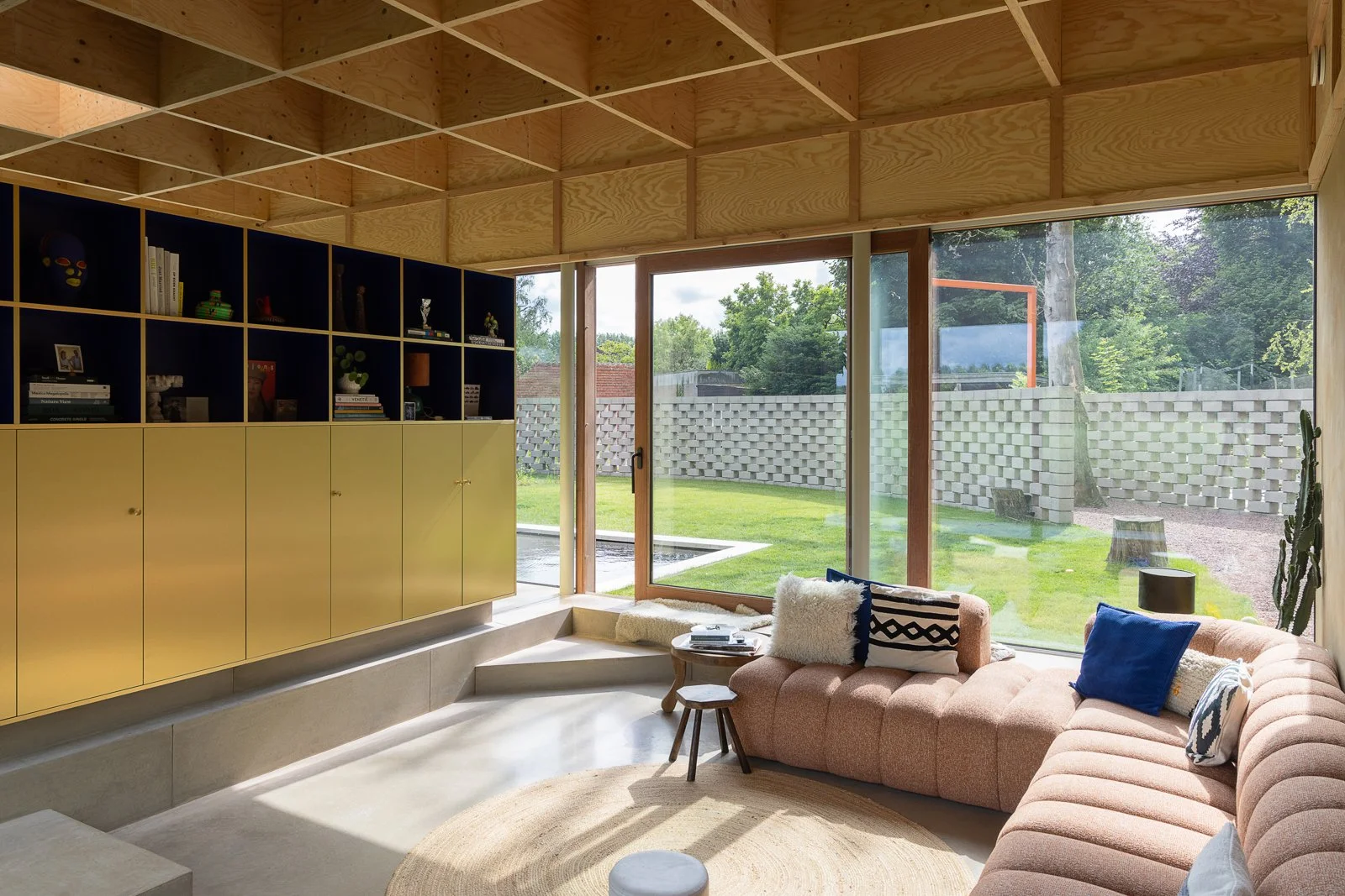 Living room with large glass sliding doors showing a backyard with green grass, trees, and a concrete block wall. The room has a pink sectional sofa with pillows, a small wooden side table, a round rug, and a black shelving unit with closed cabinets 
