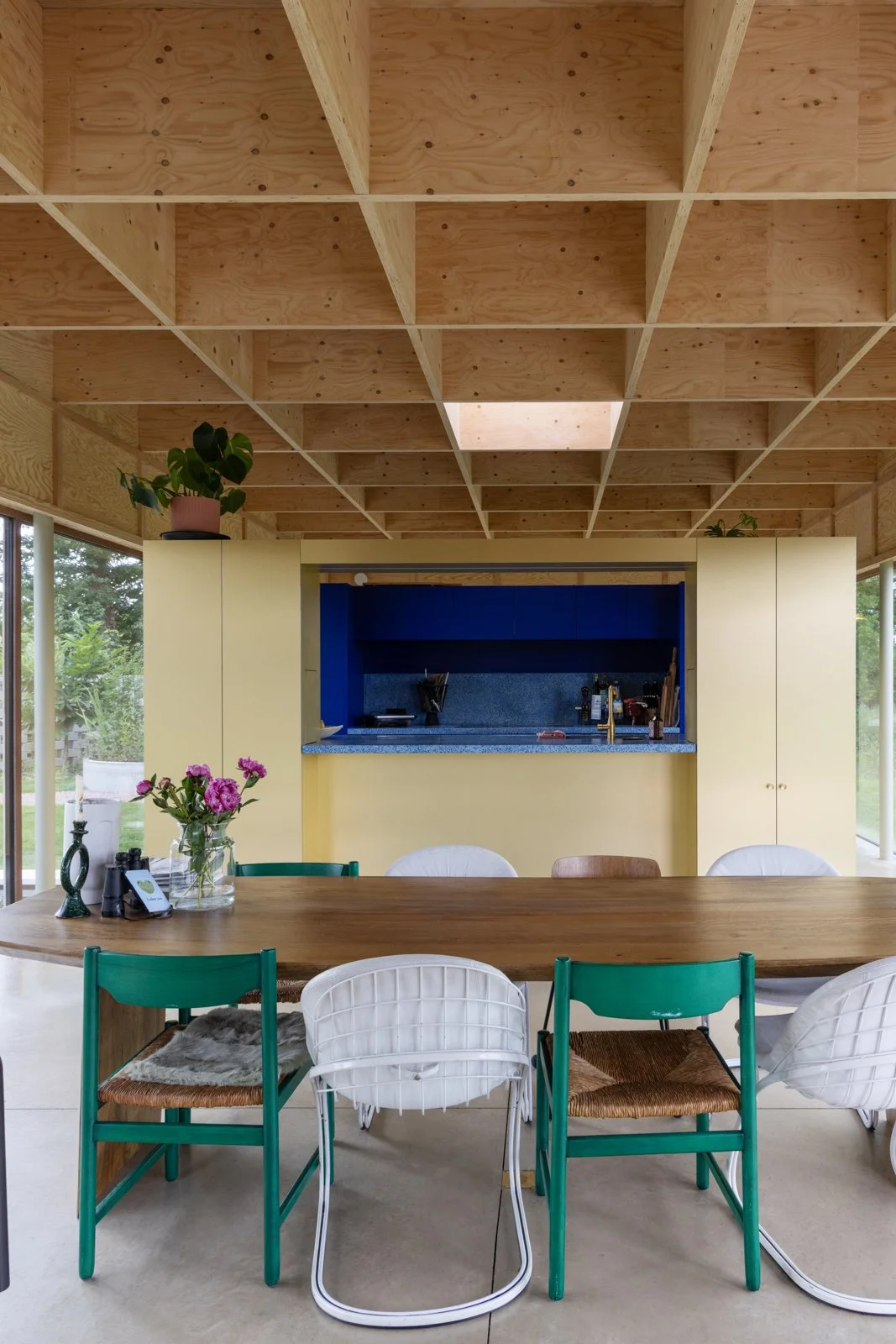 Interior of a modern kitchen and dining area with a wooden ceiling, a large wooden table, and various chairs. There are plants on top of cabinets, and an outdoor garden is visible through large glass sliding doors.