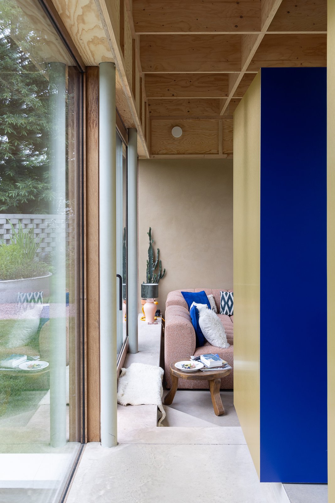 Living room corner with pink sofa, blue and white pillows, wooden side table with books, white shaggy rug, potted cacti, large glass sliding door, and wooden ceiling.