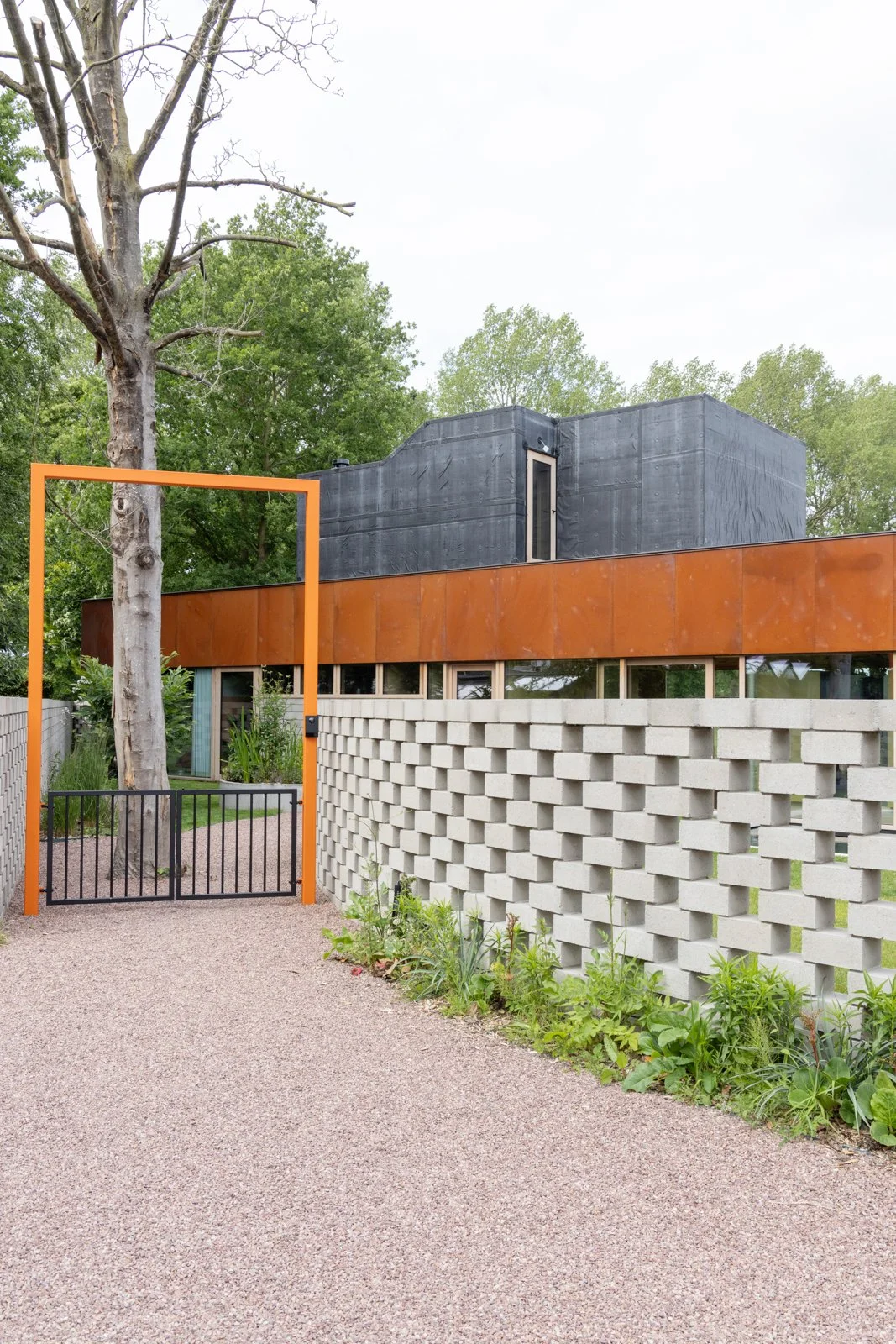 Modern house with geometric design, featuring a textured concrete block wall, a rust-colored metal band, and black upper sections, set behind an orange frame gate and surrounded by greenery.