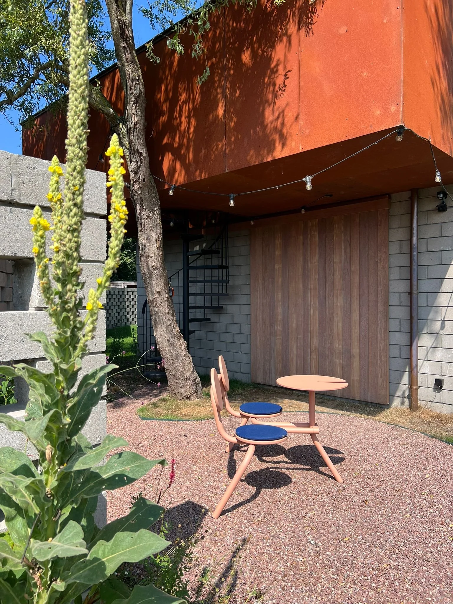 Outdoor patio with two peach-colored chairs with blue cushions and a matching small round table on a pebble surface. A tree and yellow-flowered plant are in the foreground. The background features a building with exposed brick, wood paneling, and a m