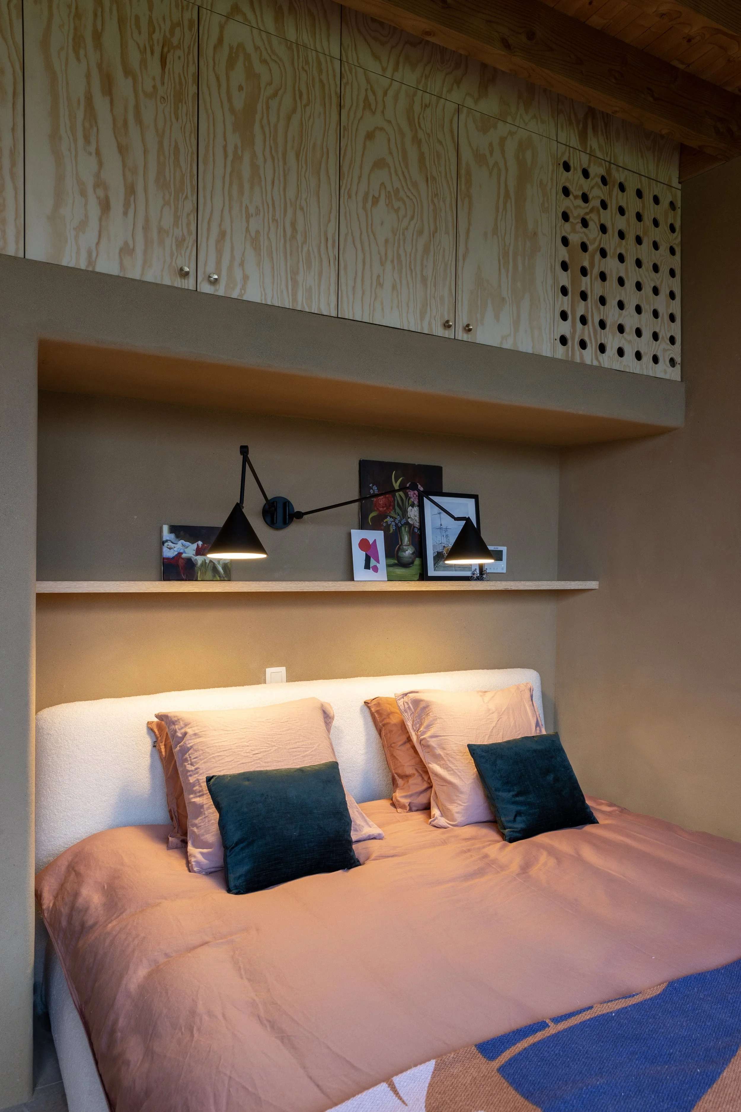 A cozy bedroom featuring a bed with peach-colored bedding and four pillows, two in peach and two in dark green, against a beige wall with built-in shelves and artwork, with black wall-mounted reading lamps.