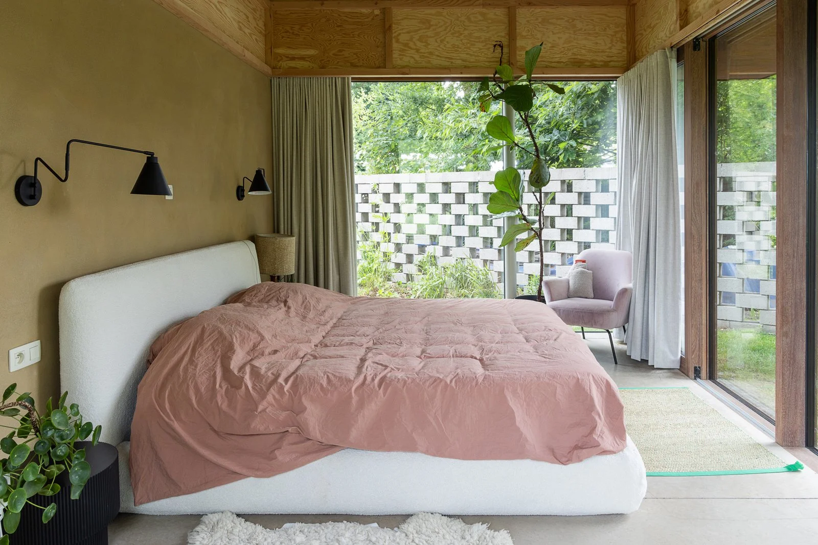 A bedroom with a large bed covered in a pinkish-brown blanket, a white headboard, black wall-mounted lamps, a nightstand with a beige lamp, a small potted plant, and a sitting area outside with a pink cushioned chair and a tall potted plant, visible 