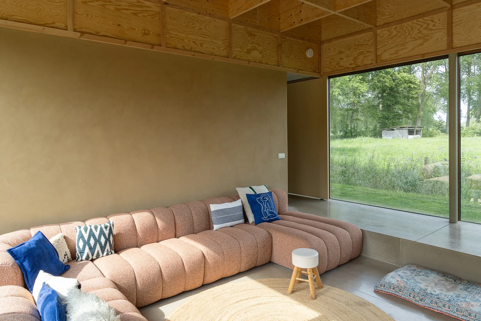 Modern living room with a large pink sectional sofa, colorful pillows, a rabbit fur throw, a round beige rug, small white stool with wooden legs, and a large floor-to-ceiling window showing green outdoor scenery, including trees and a small shed.