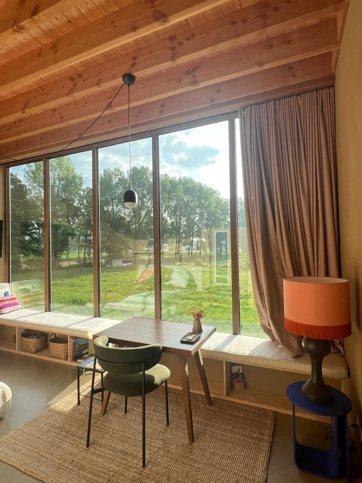 Living room with large glass window, wooden ceiling, a small wooden desk with a chair, a vase with flowers, a wooden built-in bench with cushions and baskets, and a blue side table with a lamp, overlooking greenery outside.