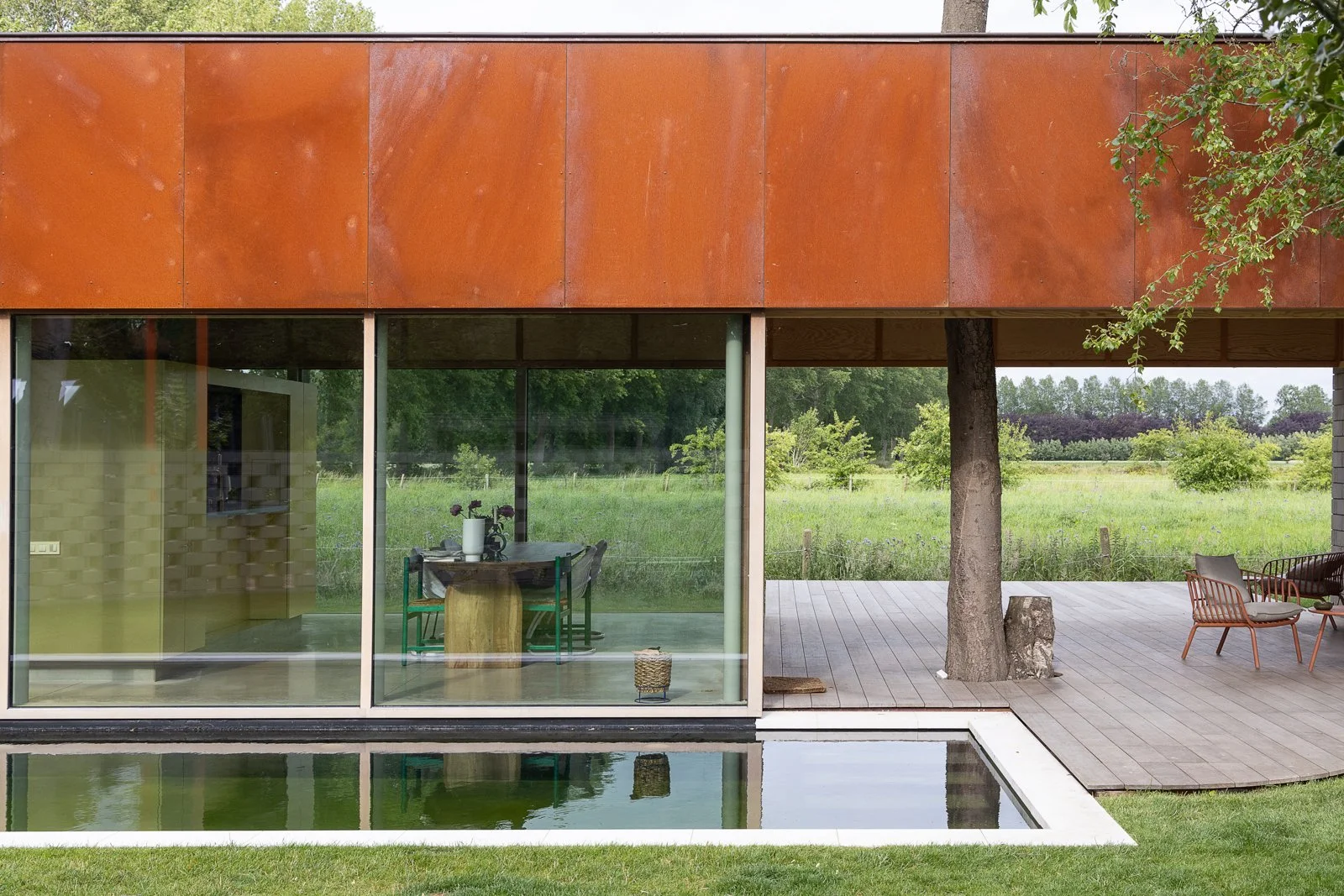 Modern house with large glass windows, a small reflecting pool in the foreground, wooden deck, outdoor seating, and trees in the background.