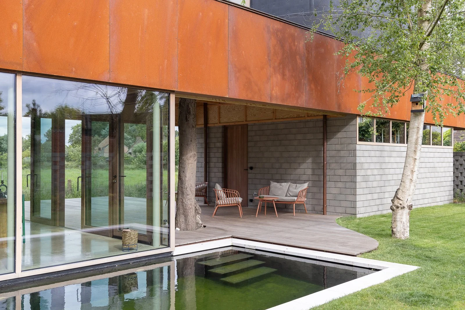 Modern house exterior with large glass windows, a wooden deck with outdoor furniture, trees, and a water feature with small steps.
