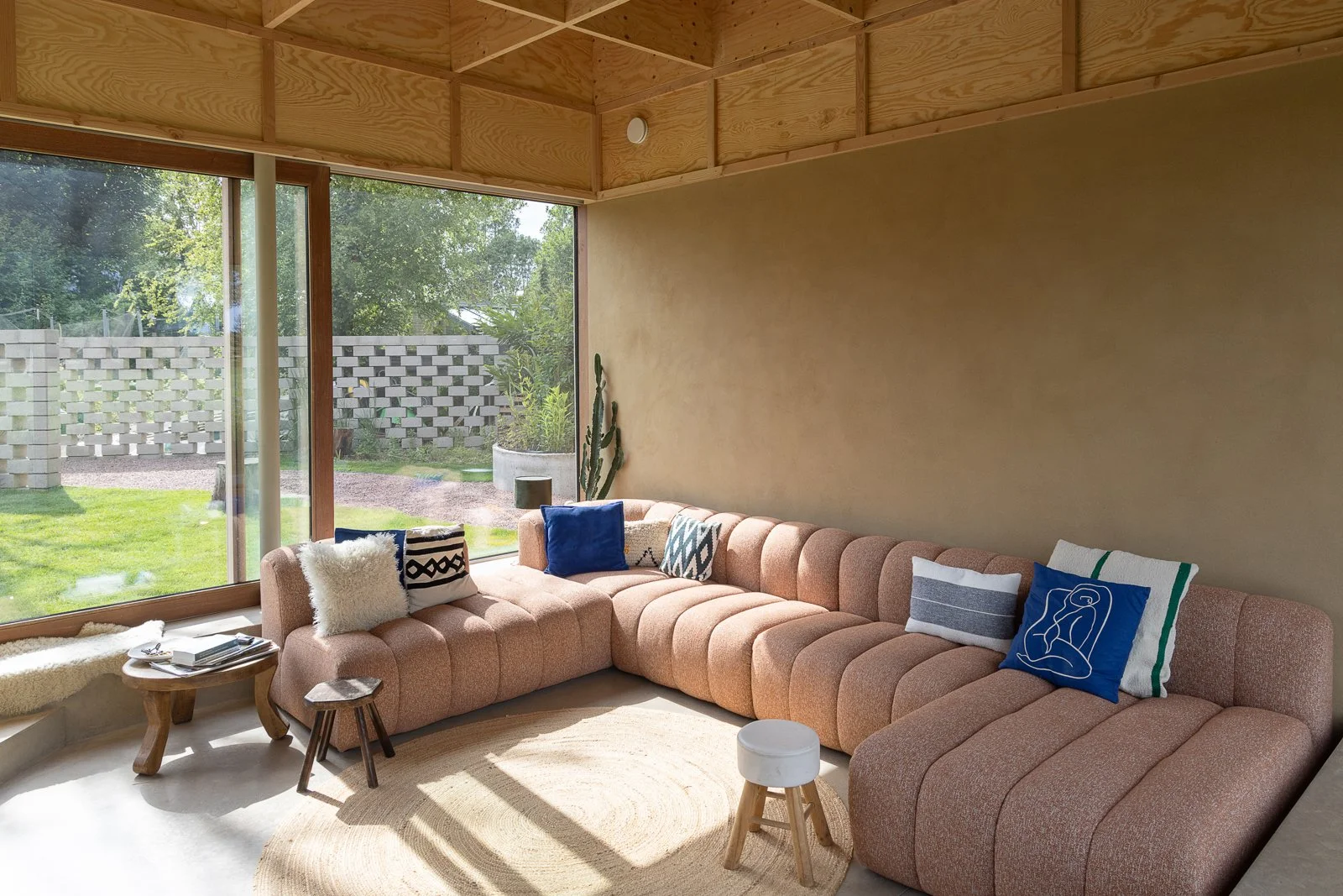 A modern living room with a large sectional pink sofa decorated with various pillows, sitting near a floor-to-ceiling window that overlooks a green backyard with trees, a concrete block wall, and potted plants. There is a small wooden stool, a coffee