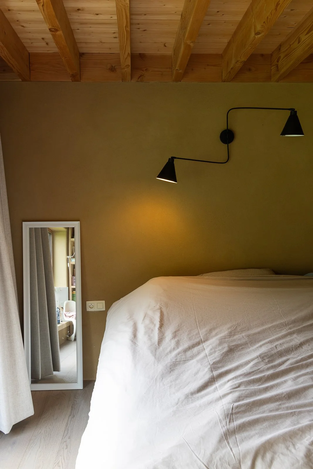 A cozy bedroom with a beige wall, a black modern wall lamp, a large bed with white bedding, a small mirror leaning against the wall, and a glimpse of a bathroom in the background.