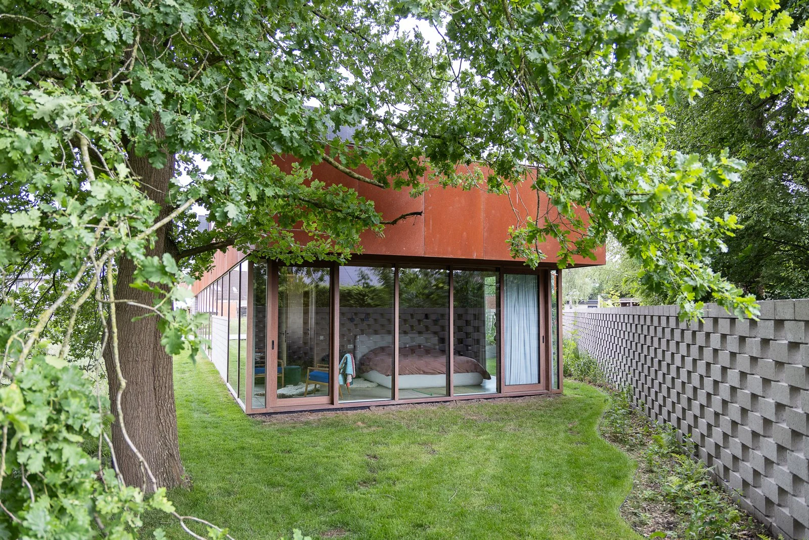 A modern house with large glass windows and a bed visible inside, surrounded by green trees and grass, with a textured concrete wall on the right.