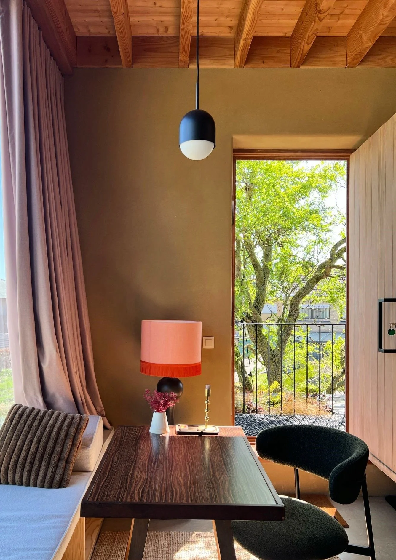Interior of a cozy room with a wooden ceiling, a black and white ceiling pendant light, a pink lampshade on a bedside table, an open door leading to a balcony with a view of green trees, and a dark green chair beside a wooden table.