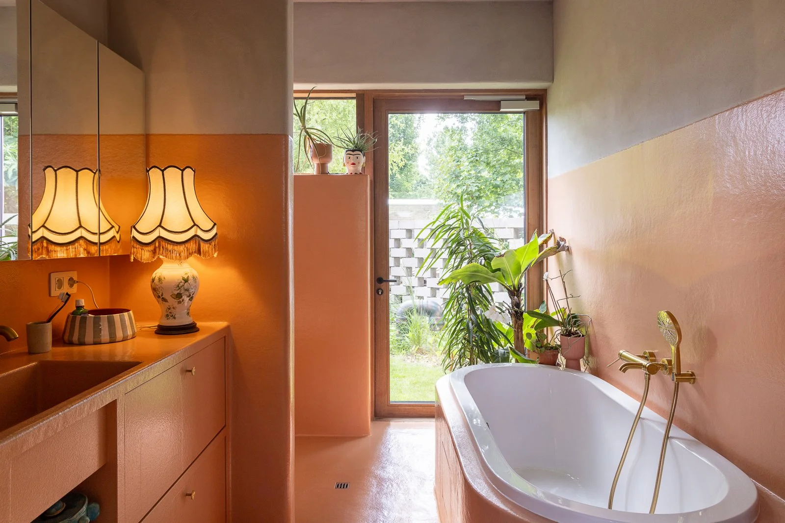 Bathroom with peach-colored walls, a white bathtub surrounded by plants, a wooden door with a glass window leading outside, and a wooden cabinet with a lampshade and decorative items.