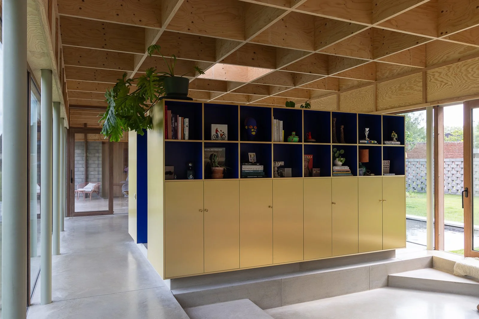 Modern interior space with a yellow and dark blue bookshelf, a potted plant on top, large glass windows, and a view of a garden outside.