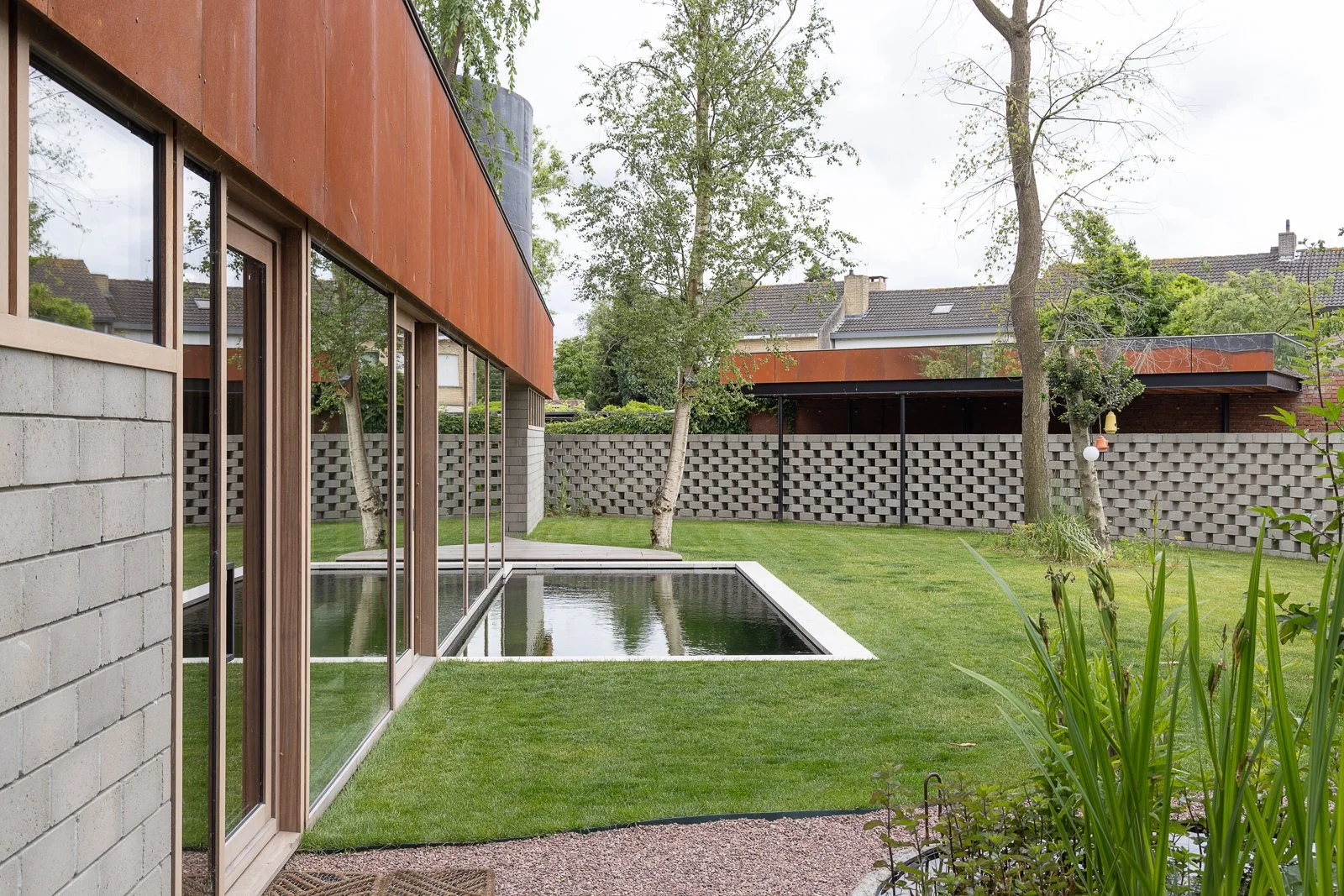 A backyard with a modern house featuring large glass windows, a swimming pool, green lawn, trees, and a patterned concrete block fence.