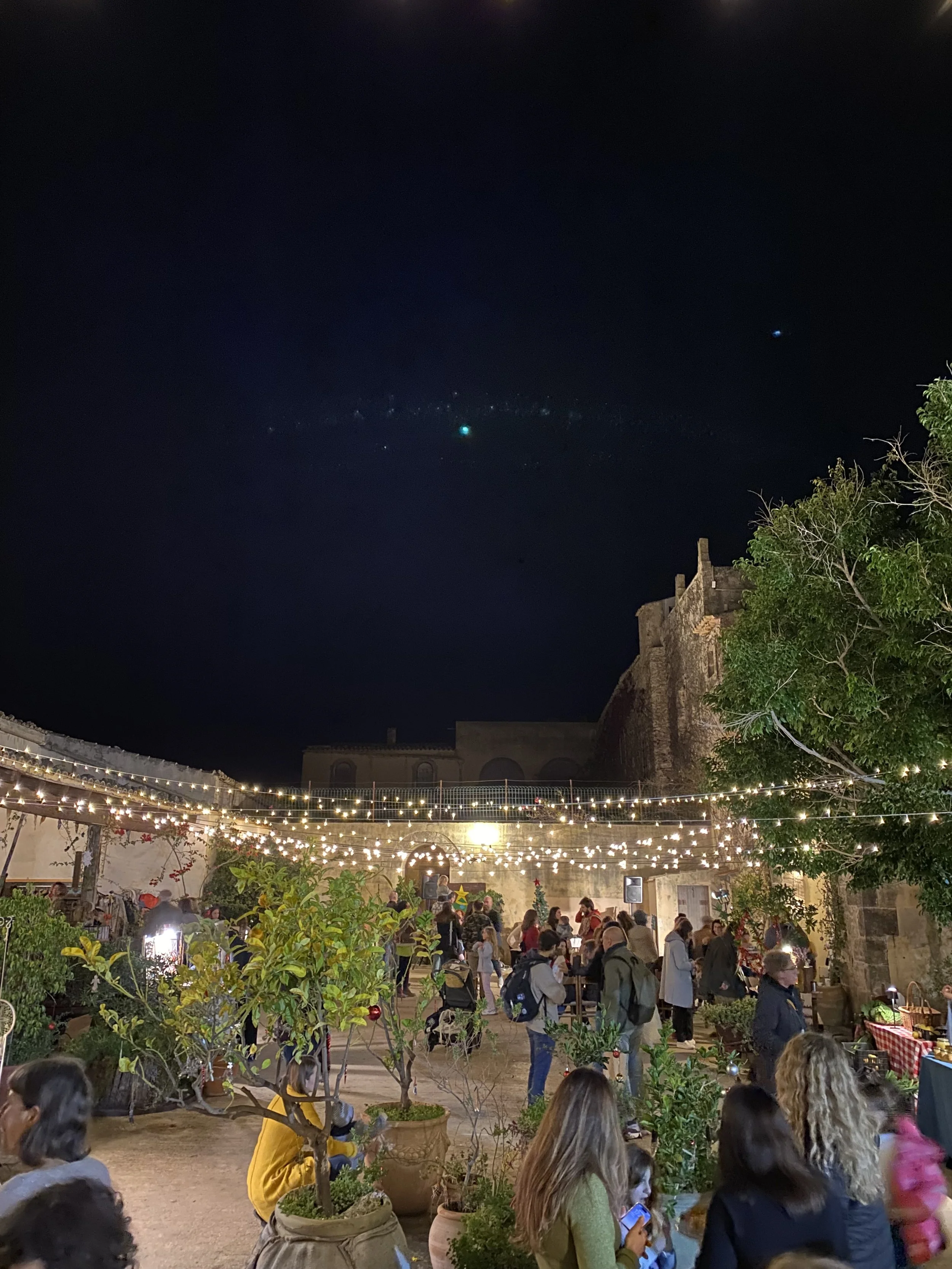 Outdoor night scene with string lights hanging above a crowd of people, some seated and others standing, at a festive gathering. The background shows an old stone building and part of a fortress wall with trees nearby.