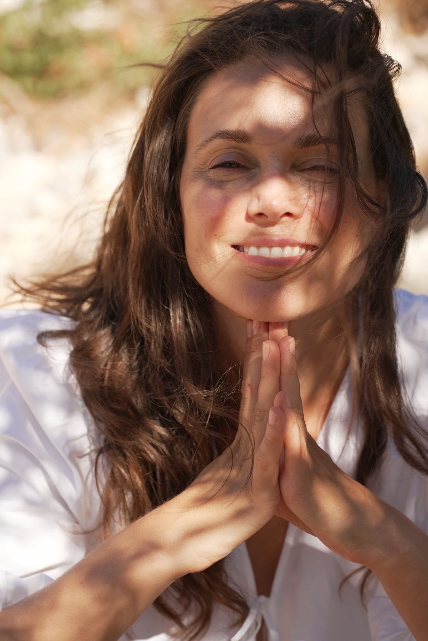 Close-up of a woman with long, wavy brown hair smiling with hands clasped together in front of her face outdoors.