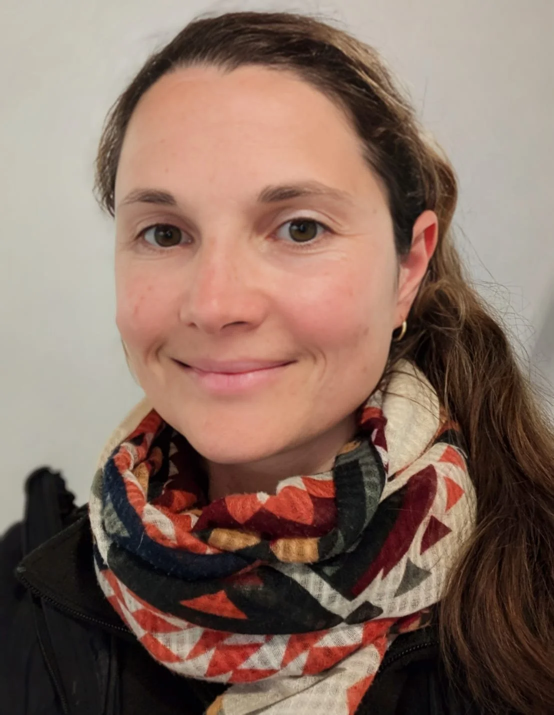 Close-up of a woman with brown hair, light skin, wearing a patterned scarf and a black jacket, smiling at the camera.