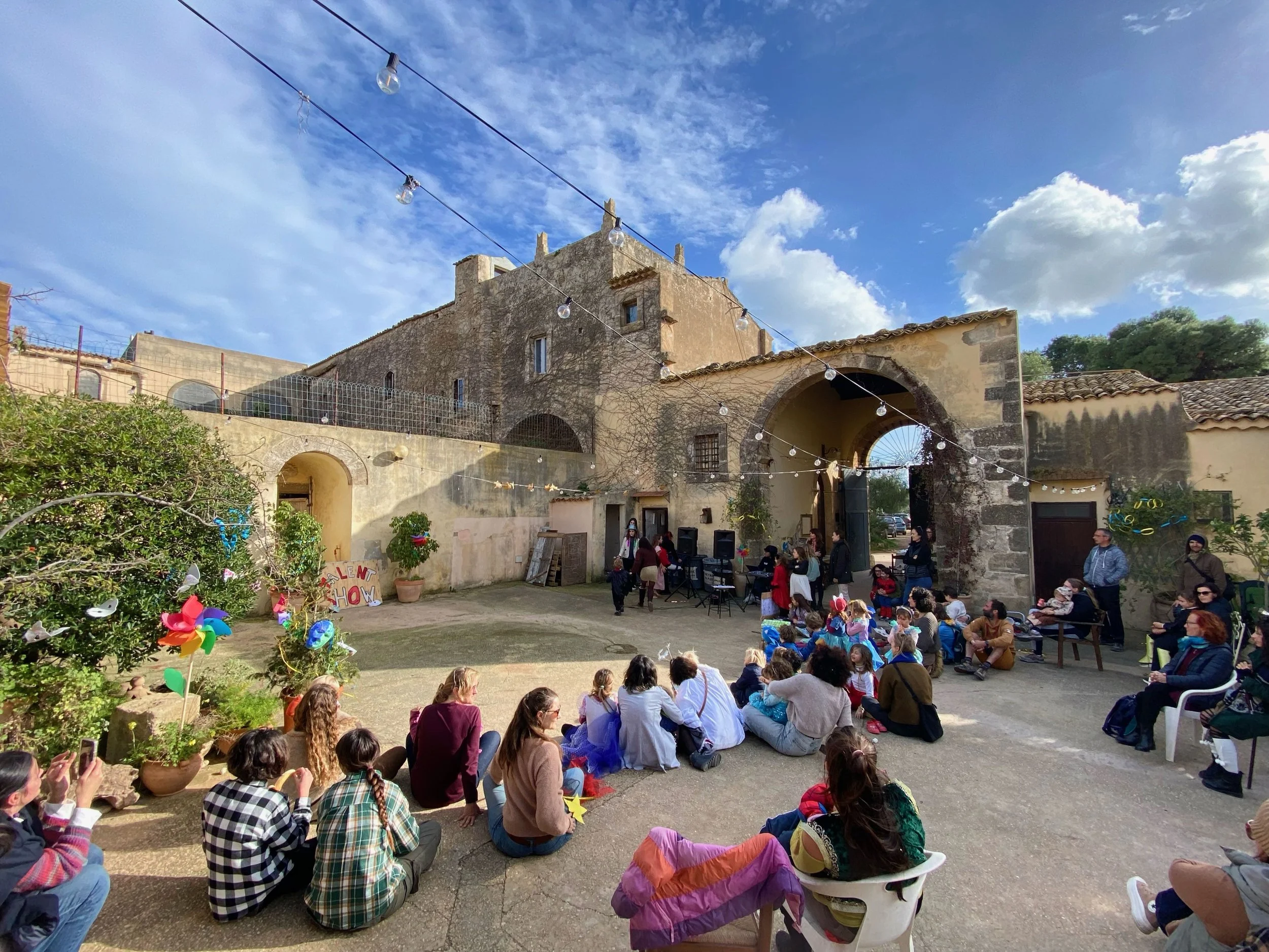 A courtyard scene with a group of children and adults seated on the ground and in chairs, watching a performance or presentation under string lights, in front of an old stone building with arched openings, on a sunny day with a blue sky and some clouds.