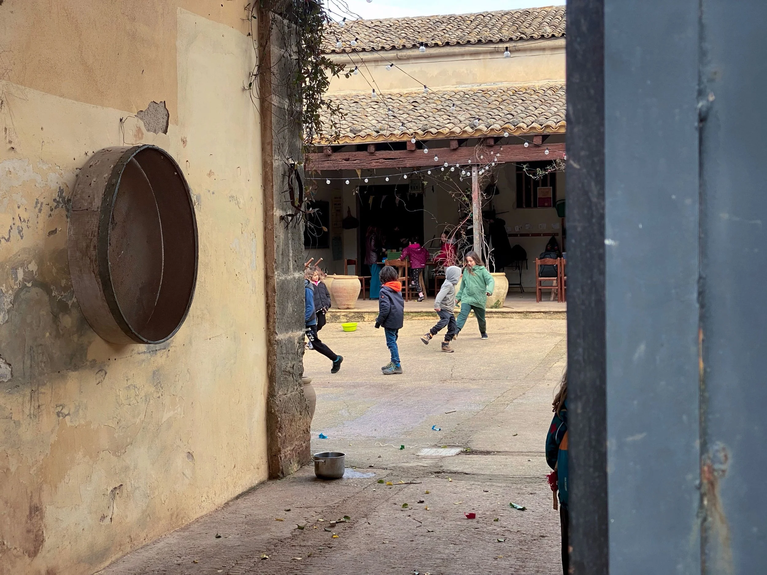 Children playing outside in a courtyard seen through a gap between a wall and a metal gate, with string lights hanging above and a building with a tiled roof in the background.