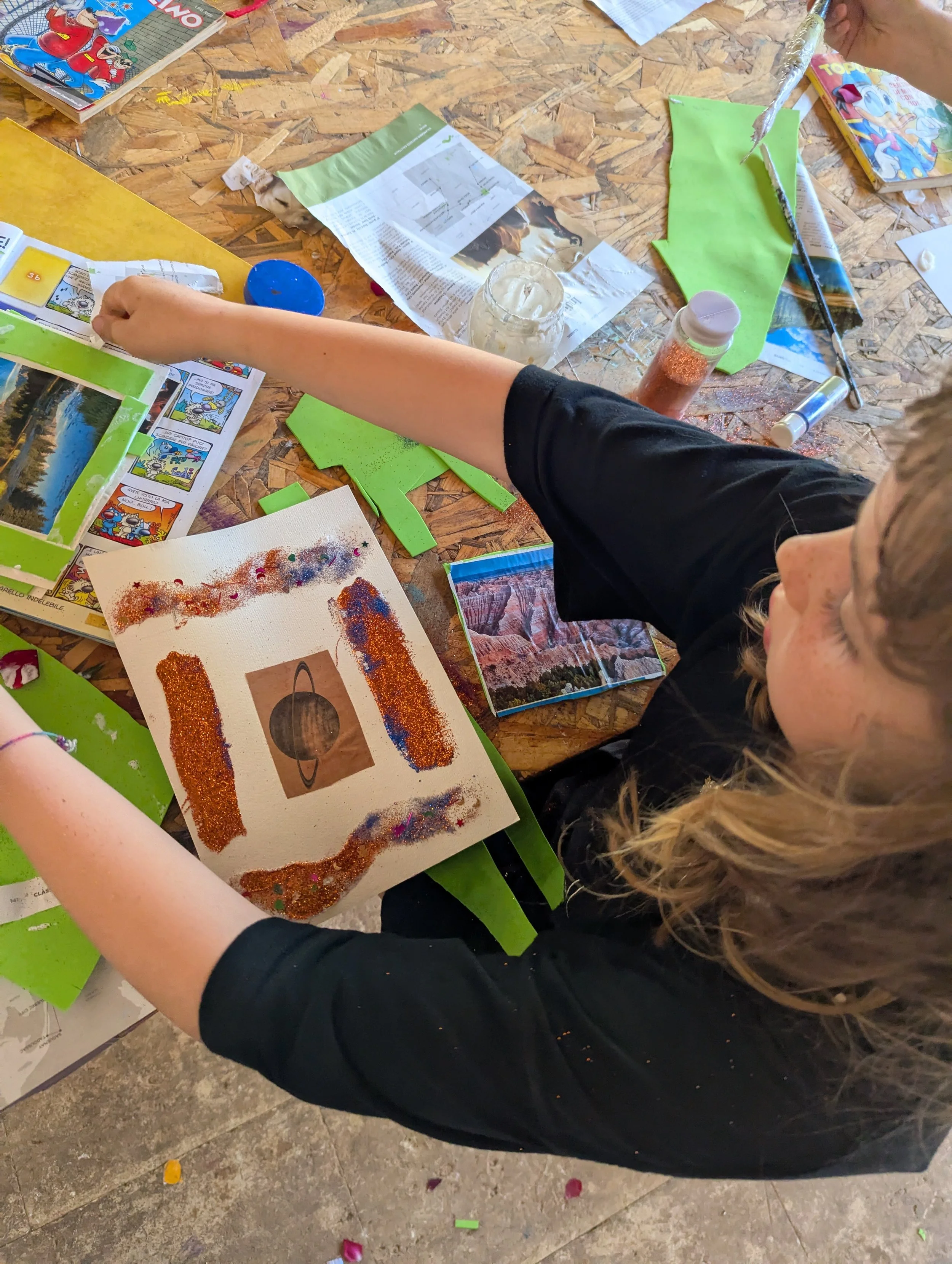 Child creating a colorful art project with glitter, paper, and photographs on a wooden table surrounded by craft supplies, magazines, and papers.