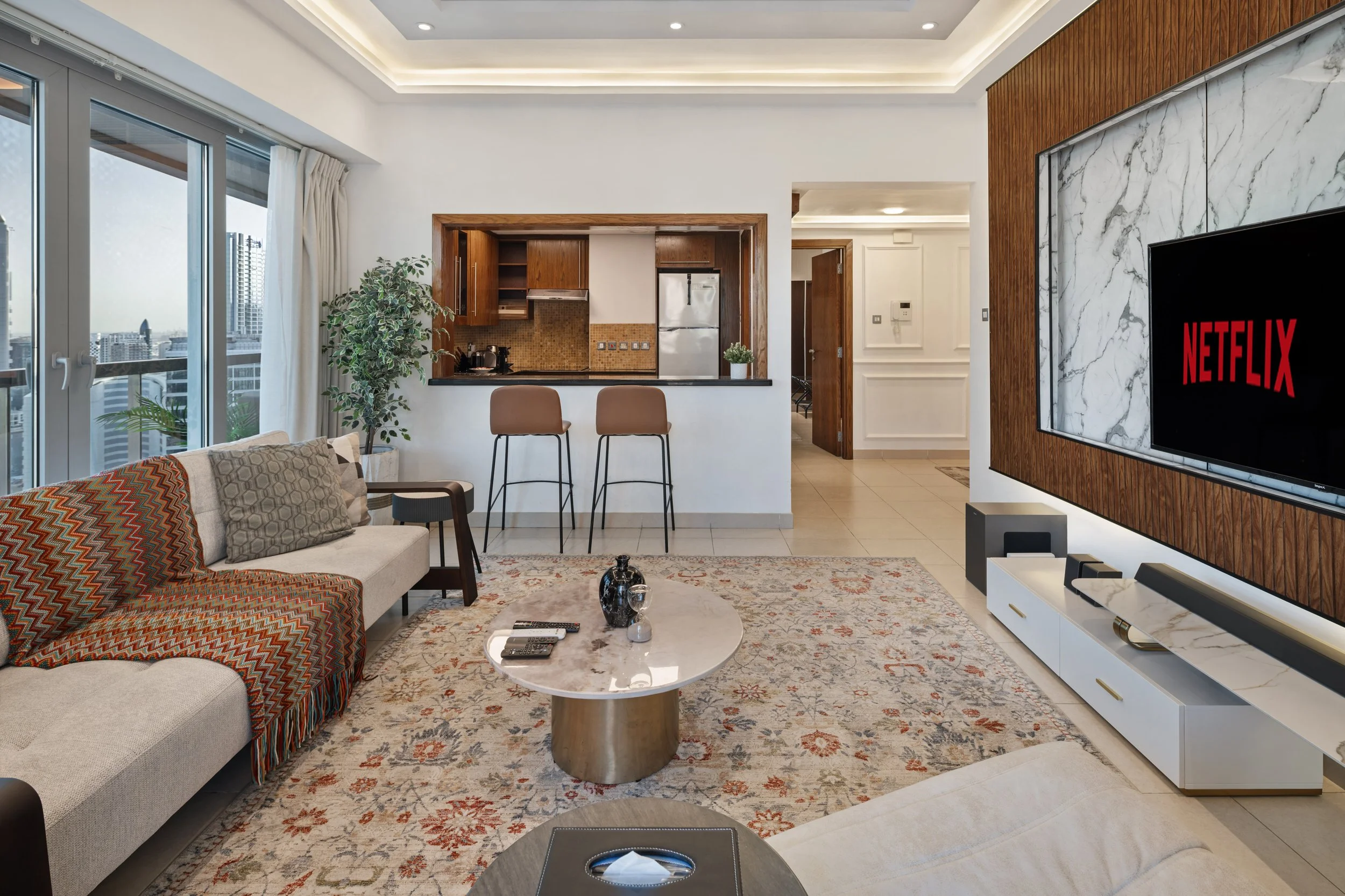 Modern living room with a large window, a patterned area rug, a beige sofa with throw pillows, a marble coffee table, a TV showing Netflix, and a view of a city outside. There is a small breakfast bar with two chairs and an open kitchen with wooden c