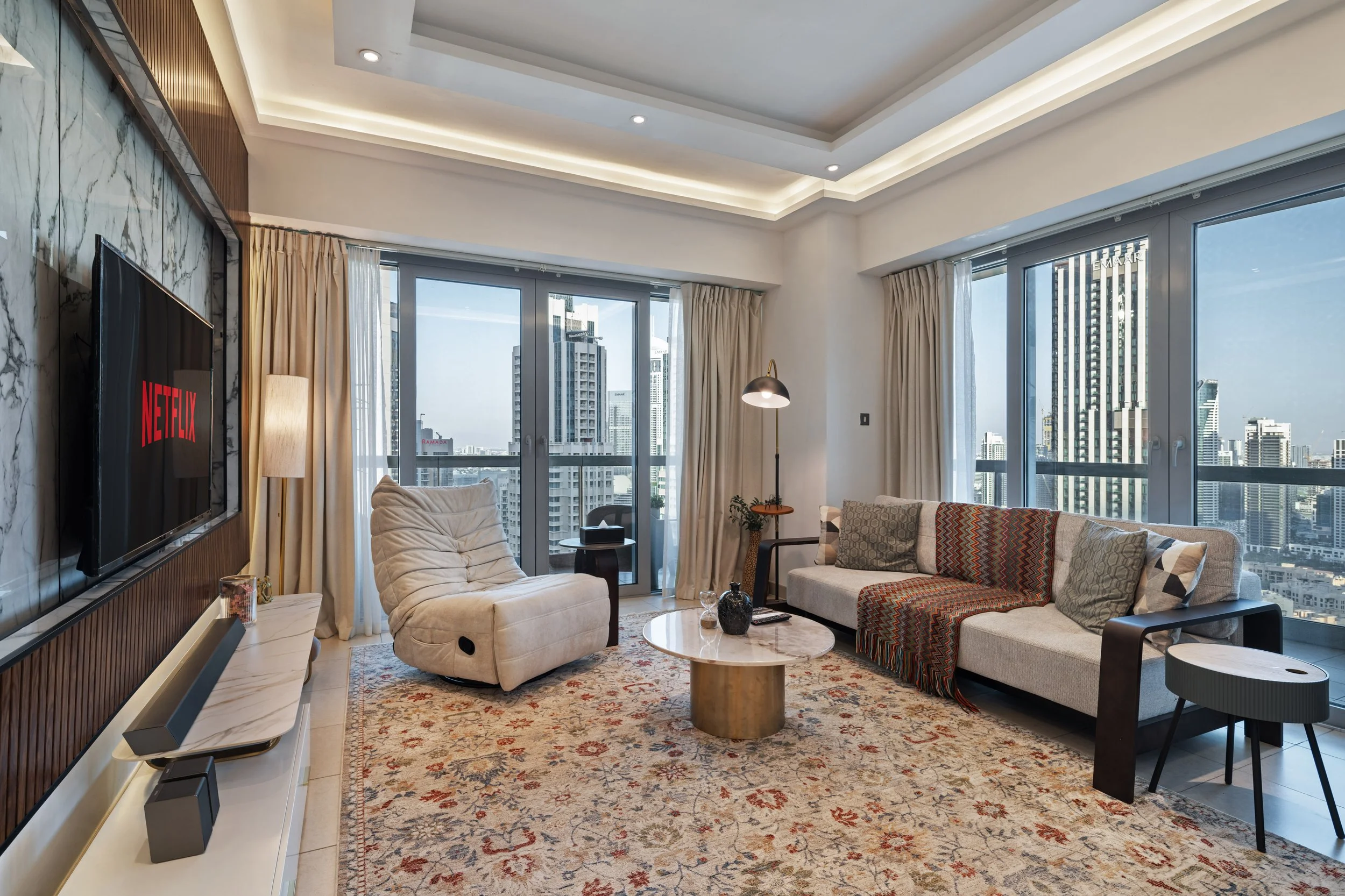Modern living room with large windows showing city skyline, beige sofa with patterned pillows, cream armchair, marble-topped coffee table, flat-screen TV displaying Netflix, and decorative rug.