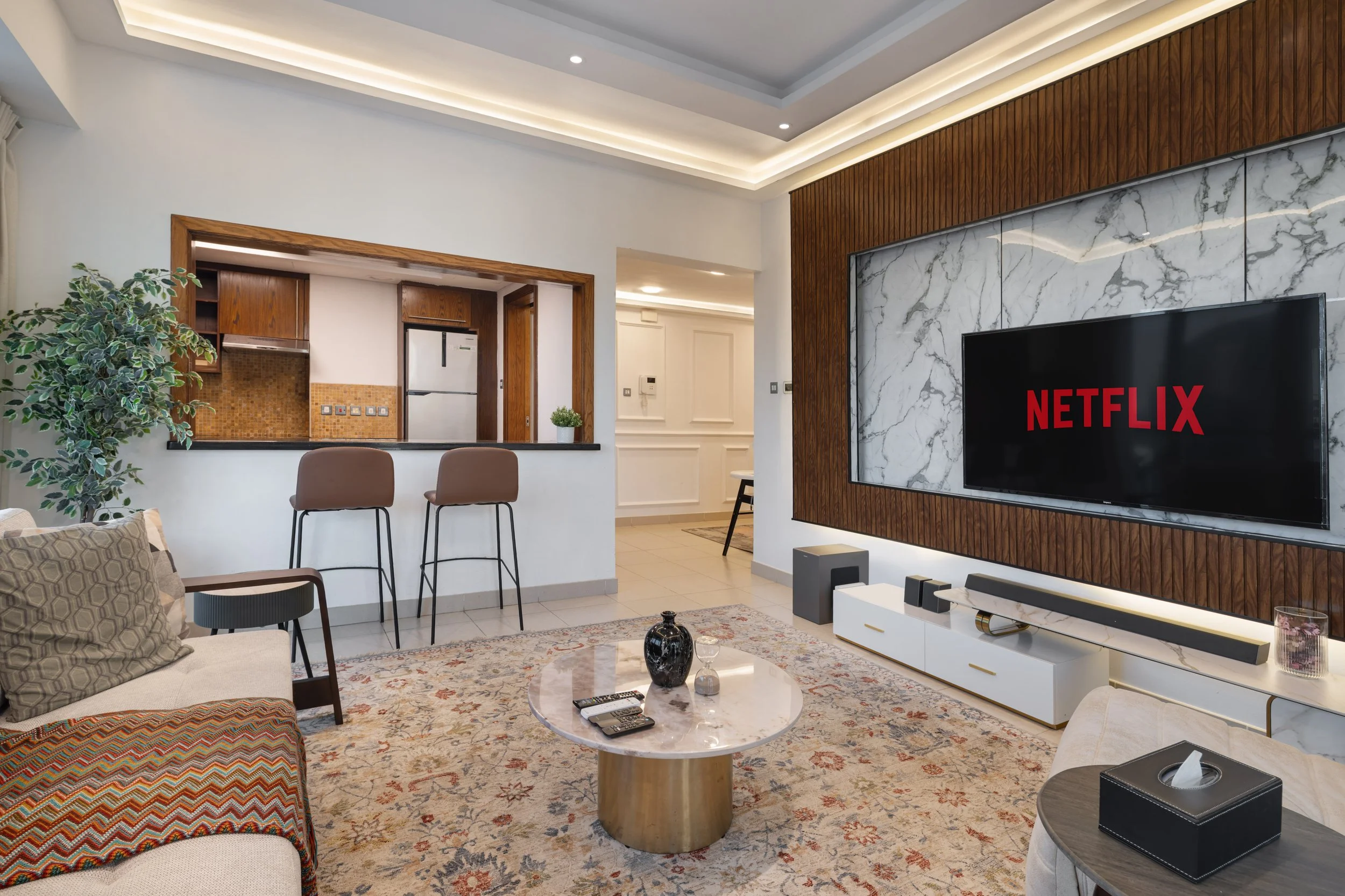 Modern living room with a TV showing Netflix, a marble and wood accent wall, a coffee table with decorative items, and a kitchen bar with stools in an open-plan design.