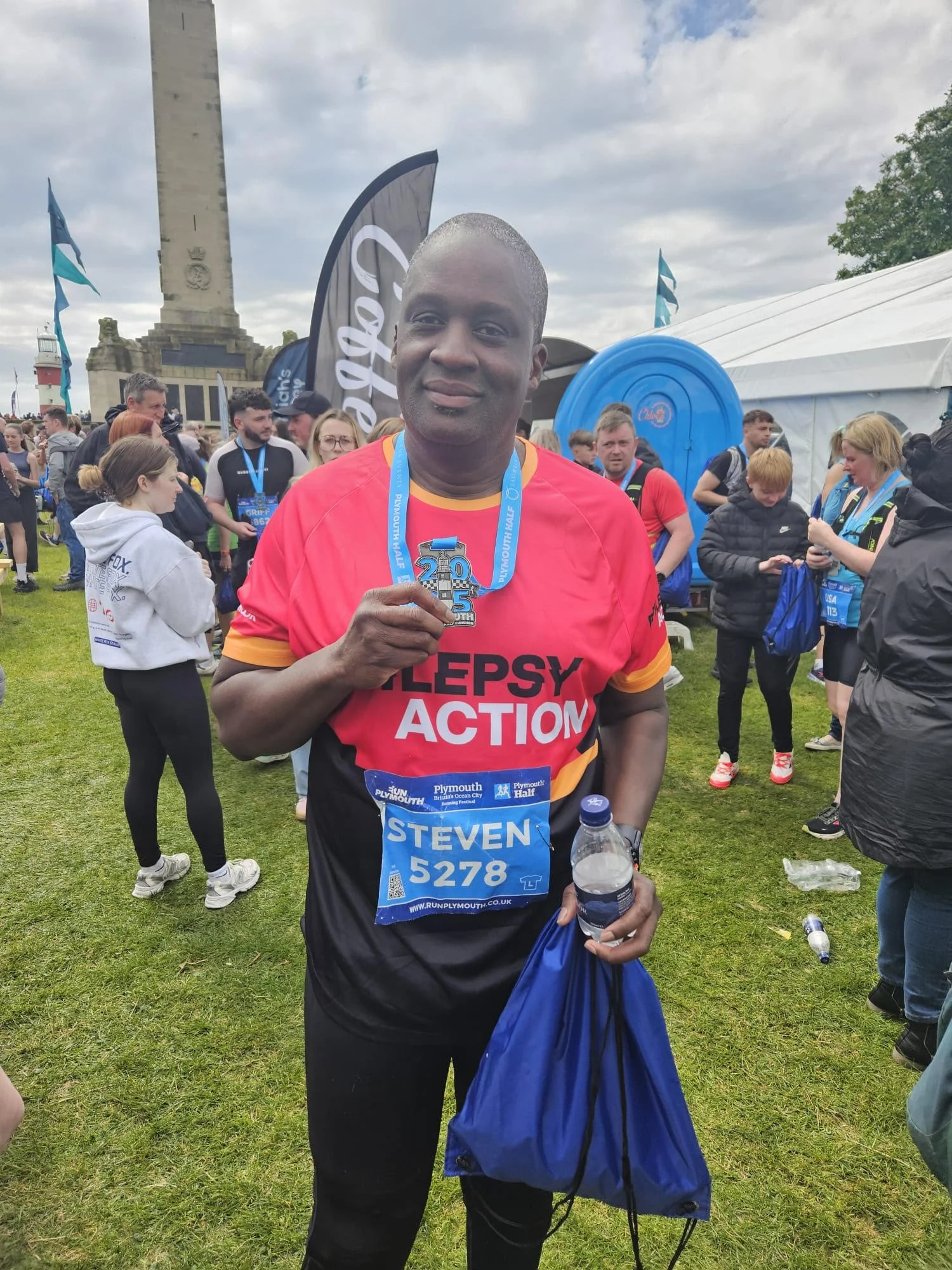 A man in a red and black running shirt holding a medal and water bottle at a race event.