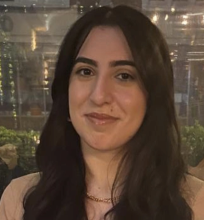 A woman with long dark hair smiling at the camera, indoors with lights and plants in the background.