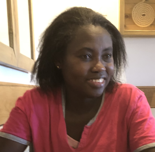 A woman with dark skin and shoulder-length black hair smiling, wearing a red shirt, sitting in a restaurant or cafe.