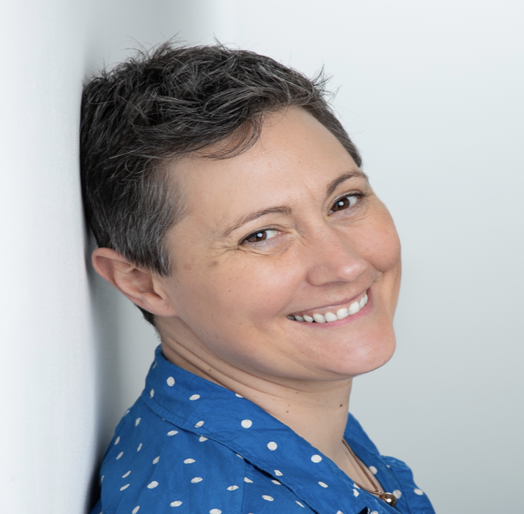 A woman with short, dark hair smiling and leaning against a white wall, wearing a blue shirt with white polka dots.