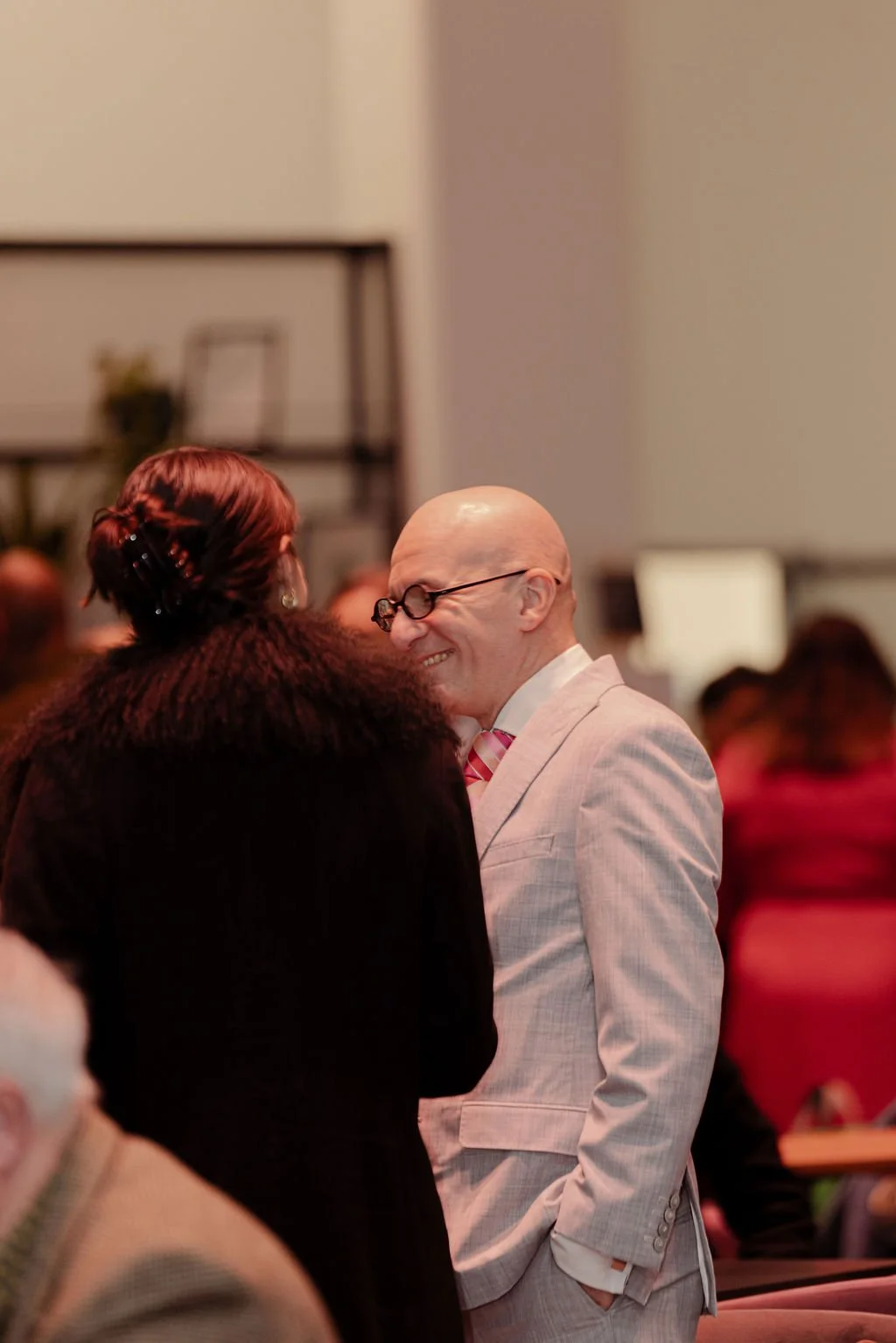 A man in a light-colored suit, pink striped tie, glasses, and a bald head smiling and interacting with a woman with dark curly hair, styled with hair clips, in an indoor setting with several other people in the background.
