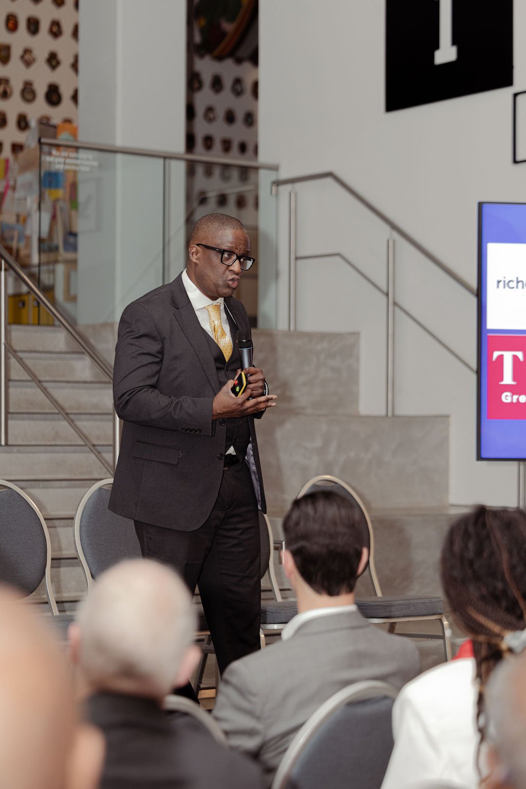 A man in a dark suit and glasses giving a presentation in a conference room, holding a microphone and a remote control, with seated audience members in front of him.