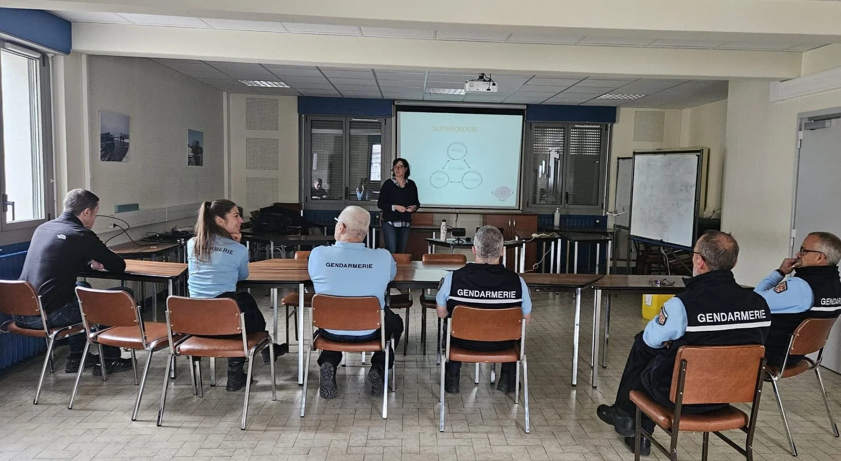 Une salle de classe avec une femme donnant une présentation sur un tableau blanc interactif, et plusieurs personnes, dont des agents de gendarmerie, assises à des tables, écoutant attentivement.