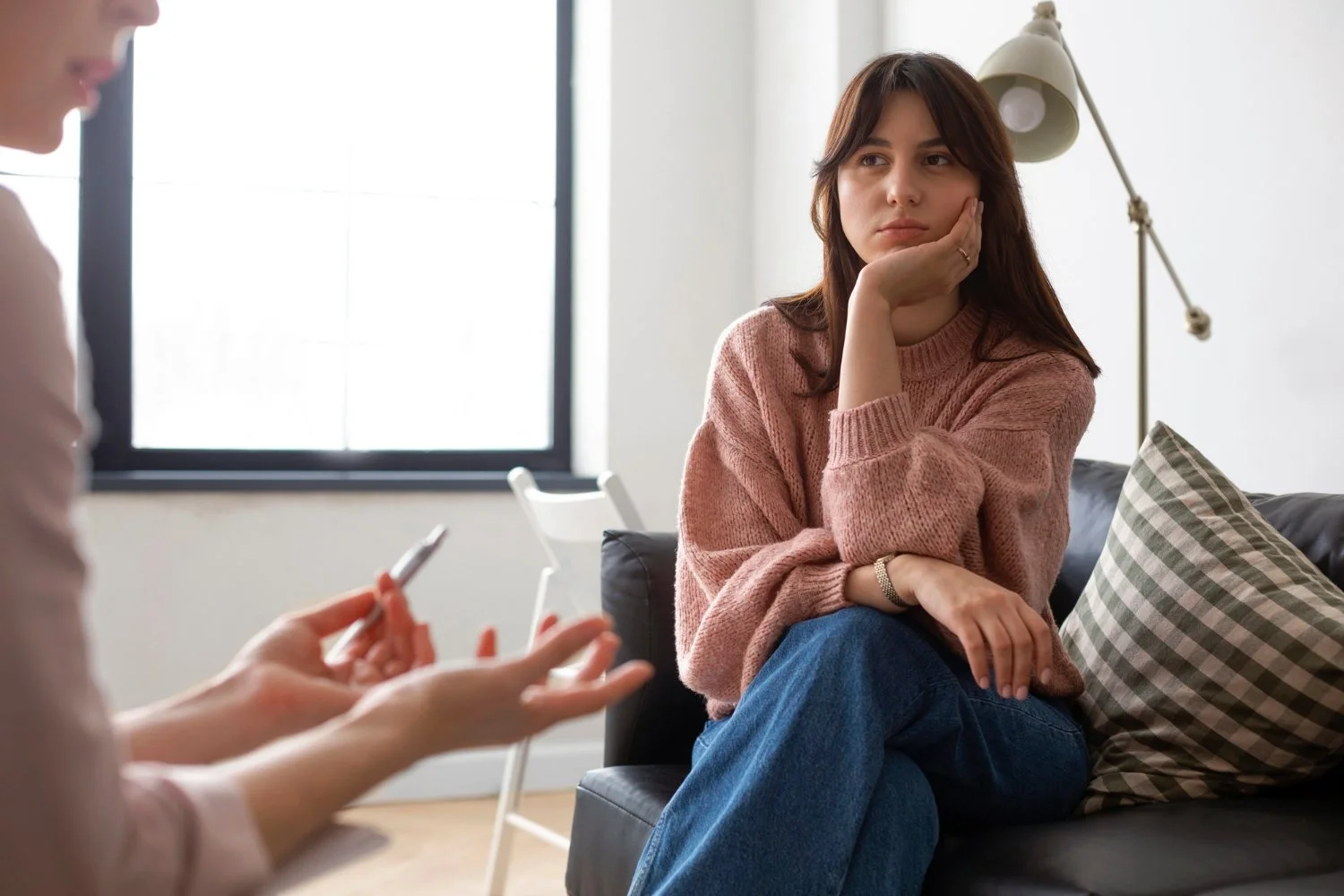 Jeune femme assise sur un canapé, semble préoccupée, en consultation avec un professionnel ou thérapeute dans une pièce lumineuse avec une lampe et une fenêtre.