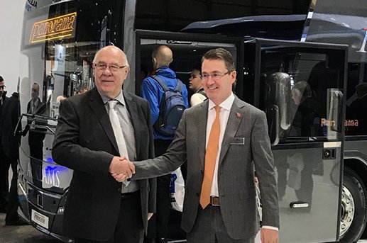 Two men in suits shaking hands in front of a bus at an indoor event.