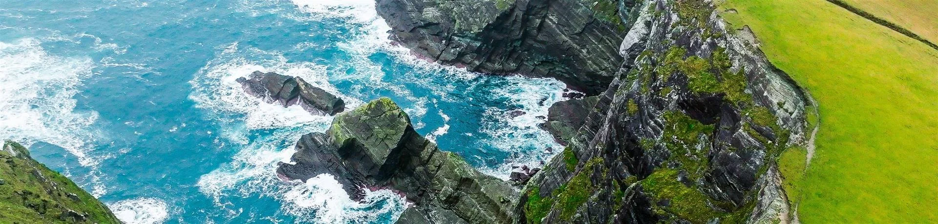 A view of a rocky coastline with waves crashing against dark cliffs and green grassy cliffs on top.