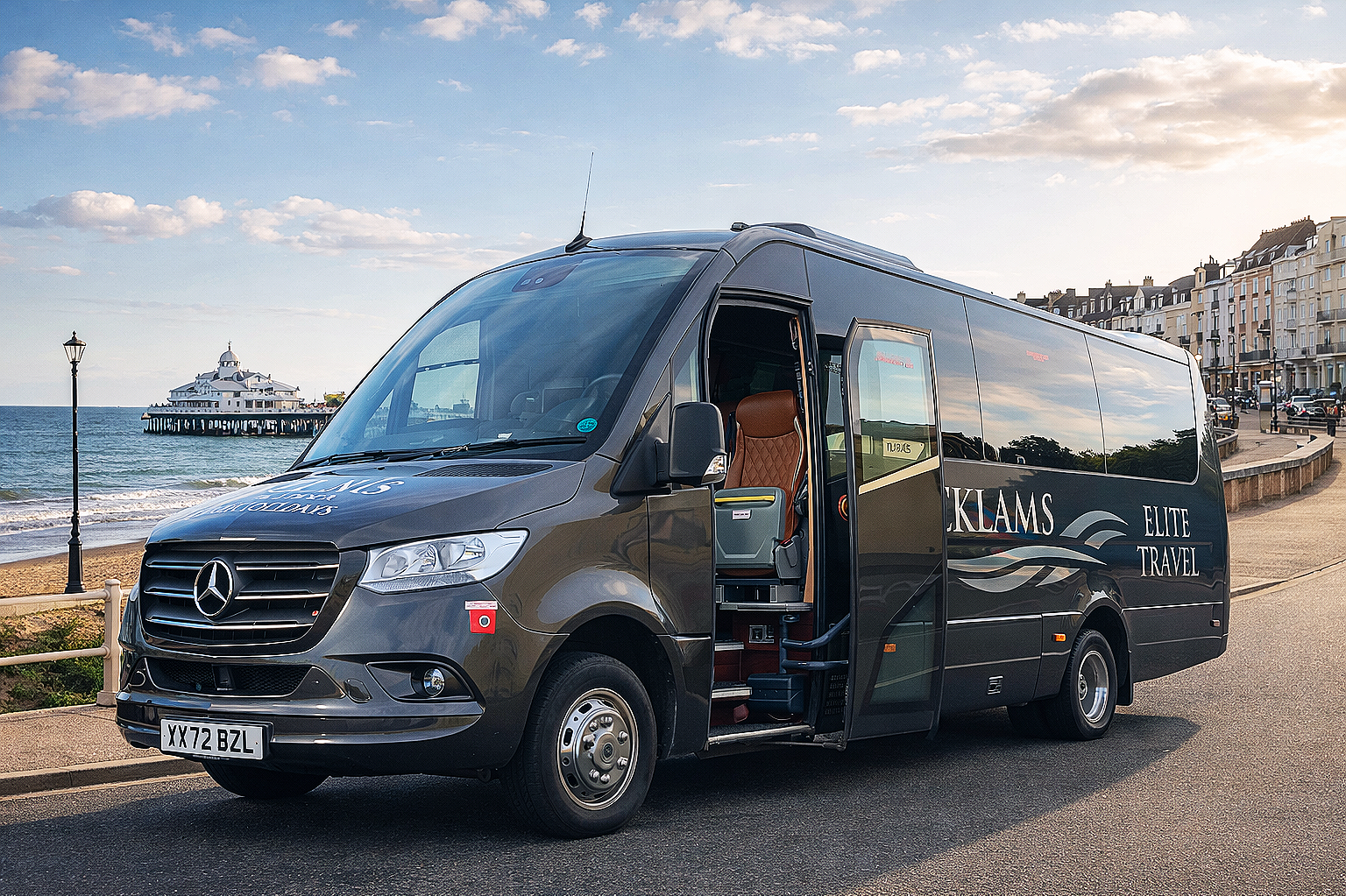 A black Mercedes-Benz travel bus parked on a road by the beach with a pier and a large building in the background, under a partly cloudy sky.