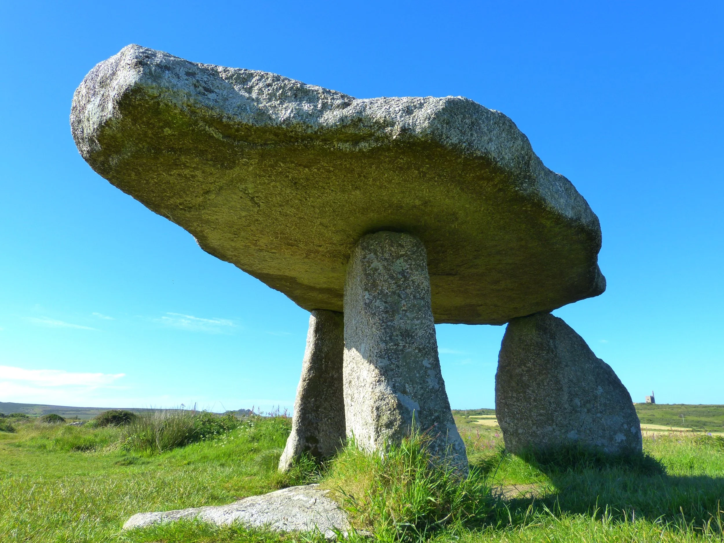 Large prehistoric stone structure known as a dolmen, with a massive flat capstone supported by three upright stones in a grassy field under clear blue sky.