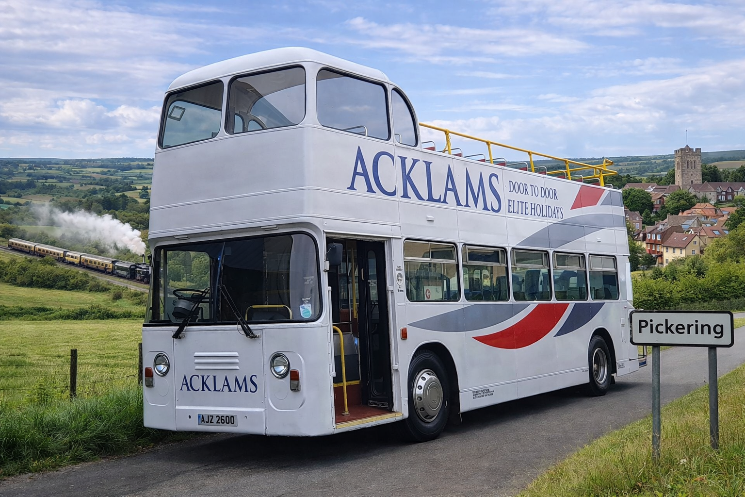 A white double-decker bus with the word "ACKLAMS" on the side, parked near a sign that says "Pickering." A steam train is visible in the background, on a hill with a village and countryside landscape.