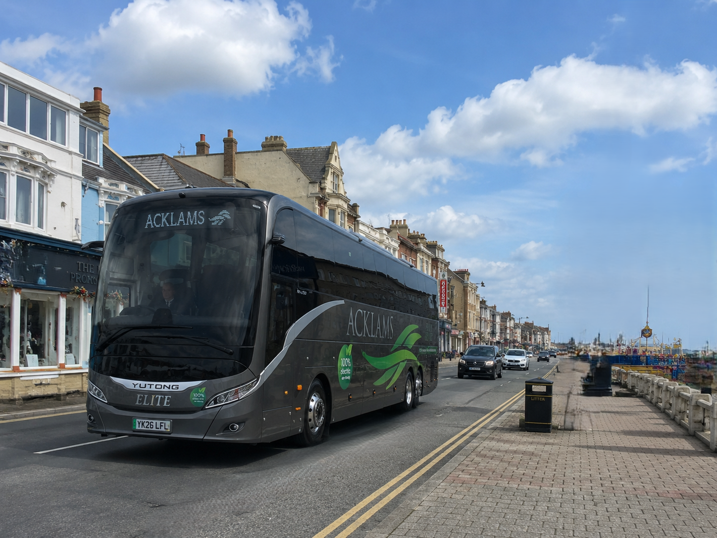 A grey electric bus on a town street with historic buildings, cars, and a playground in the background.
