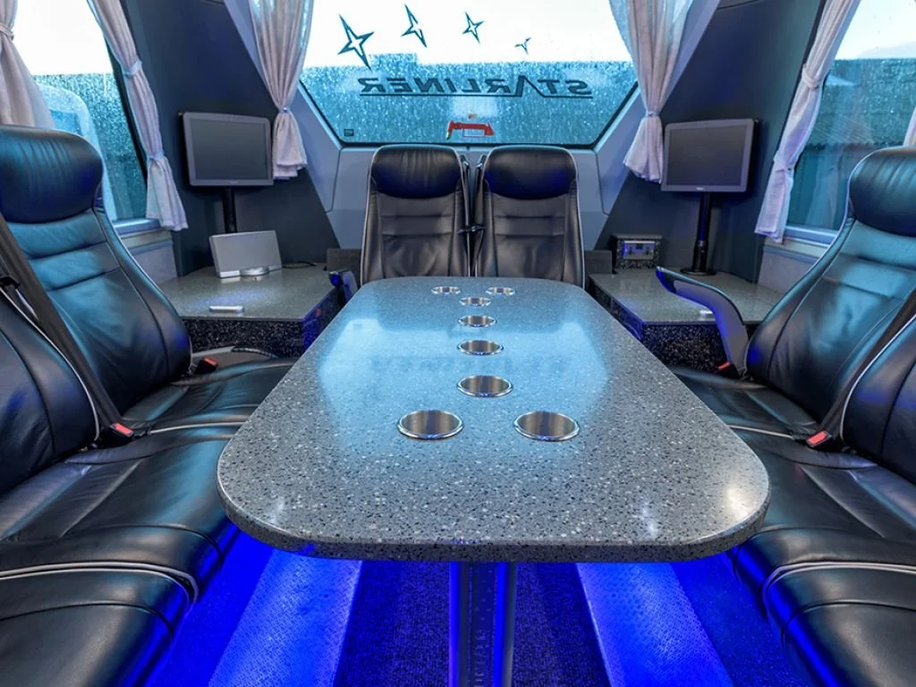 Luxury bus interior with a central conference table and black leather chairs, with two smaller screens and a window showing rain, and the word 'STARLINER' on the front window.