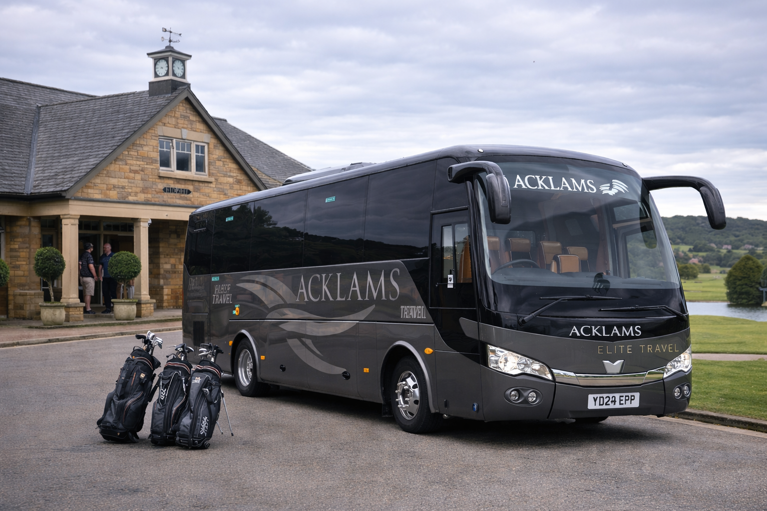 Black coach bus with 'ACKLAMS' and 'ELITE TRAVEL' branding parked in front of a stone building with a clock tower, four golf bags with clubs in the foreground, and a scenic landscape in the background.